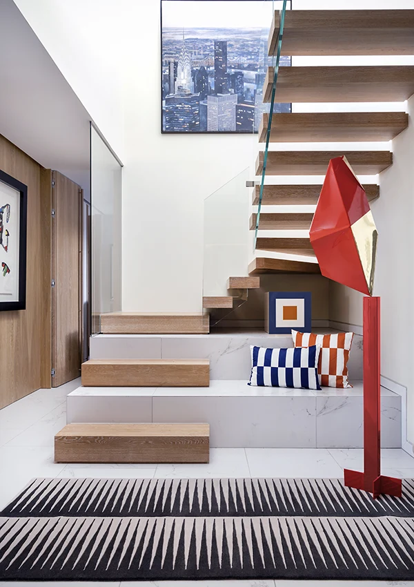 Recibidor con escaleras, decorado con cojines, alfombra y escultura con formas y estampados geométricos.