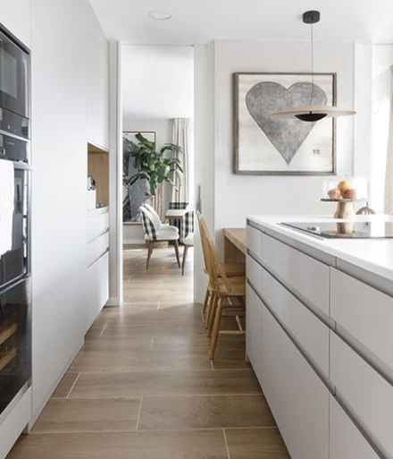 Cocina de color blanco con isla a conjunto. Mesa y sillas de madera natural a juego con el suelo.