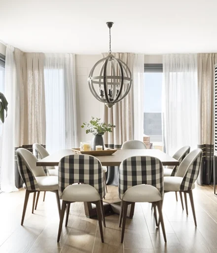 Comedor con gran mesa cuadrada y sillas en color crema, con respaldo a cuadros. Cortinas claras que permiten el paso de luz y gran lámpara colgante con forma de esfera.