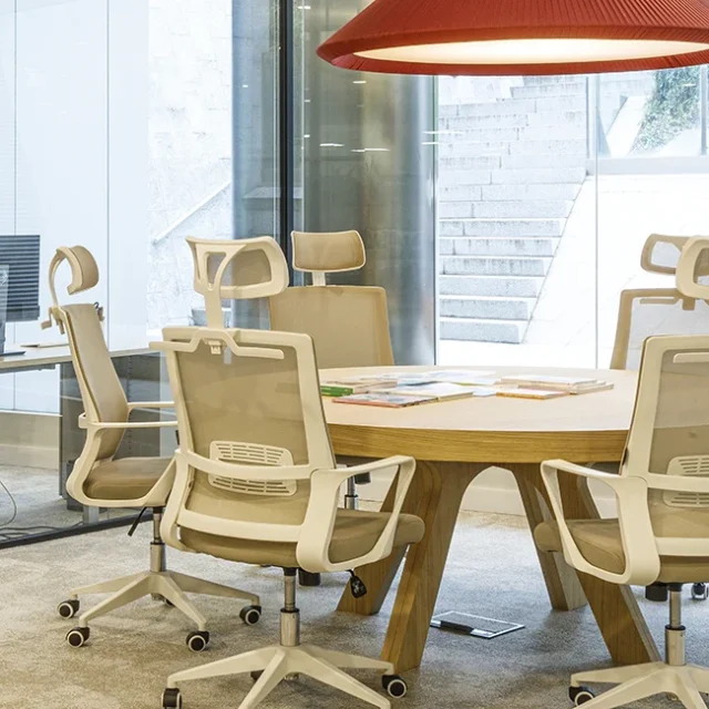 Zona de hot-desking con sillas beis y mesa redonda cerca de gran ventanal.